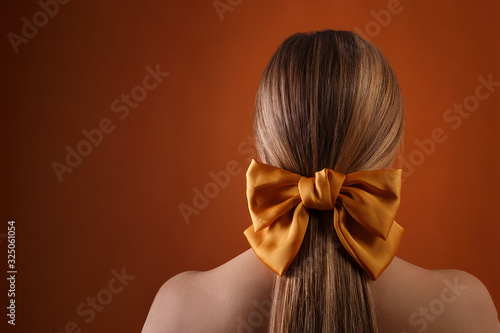 blond female hairstyle with color ribbon on orange wall background with copyspace. close up of blonde girl head with carroty bow in her hair, rear view. fashion style photoshoot of woman in studio