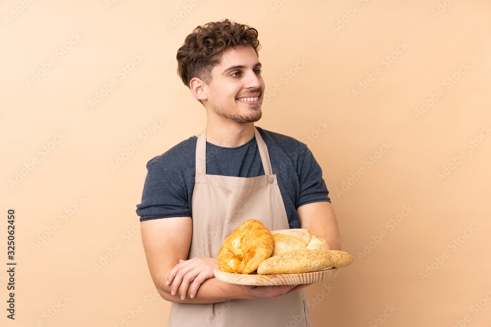 Male baker holding a table with several breads isolated on beige background looking to the side Male baker holding a table with several breads isolated on beige background looking to the side