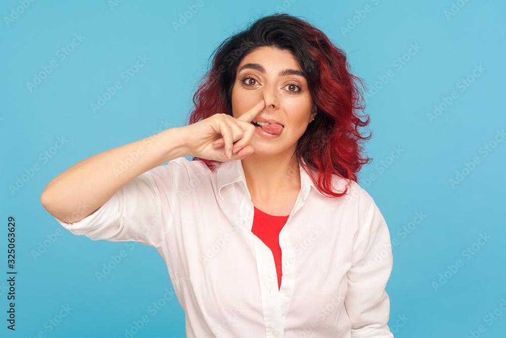 Bad manners, misconduct. Portrait of funny comical hipster woman with fancy red hair picking her nose and sticking out tongue with stupid expression. indoor studio shot isolated on blue background Bad manners, misconduct. Portrait of funny comical hipster woman with fancy red hair picking her nose and sticking out tongue with stupid expression. indoor studio shot isolated on blue background