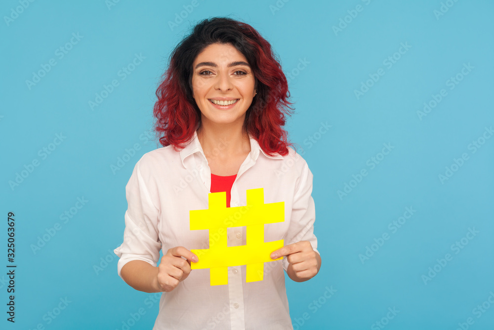 Trendy blogs. Portrait of charming woman with fancy red hair holding big hashtag symbol and smiling to camera, hash sign to tag forum discussions. indoor studio shot isolated on blue background Trendy blogs. Portrait of charming woman with fancy red hair holding big hashtag symbol and smiling to camera, hash sign to tag forum discussions. indoor studio shot isolated on blue background