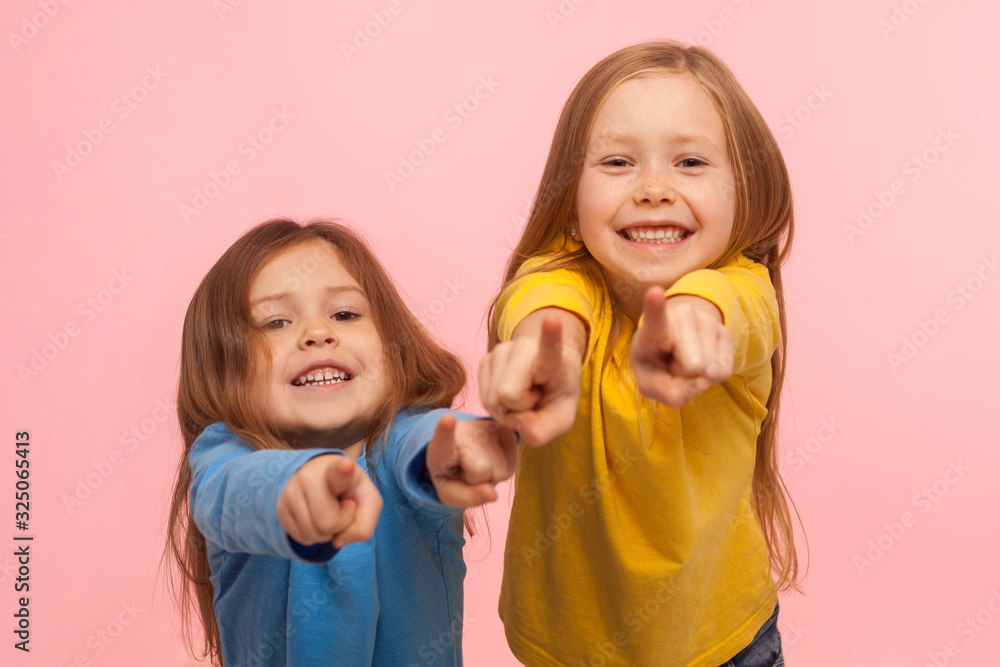 Hey you! Two charming happy little girls pointing to camera, indicating lucky one and smiling, choosing goods in child store, we need this concept. indoor studio shot isolated on pink background Hey you! Two charming happy little girls pointing to camera, indicating lucky one and smiling, choosing goods in child store, we need this concept. indoor studio shot isolated on pink background