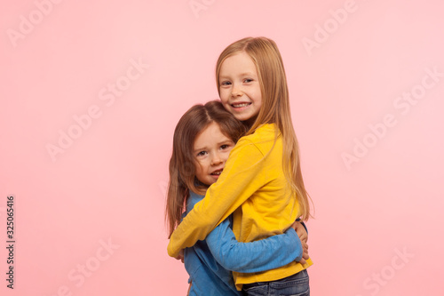 Best friends since childhood. Portrait of two adorable little girls embracing and smiling at camera with carefree happy expression, sister's support. indoor studio shot isolated on pink background