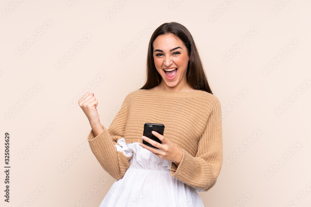 Fototapeta premium Young caucasian woman isolated on beige background with phone in victory position