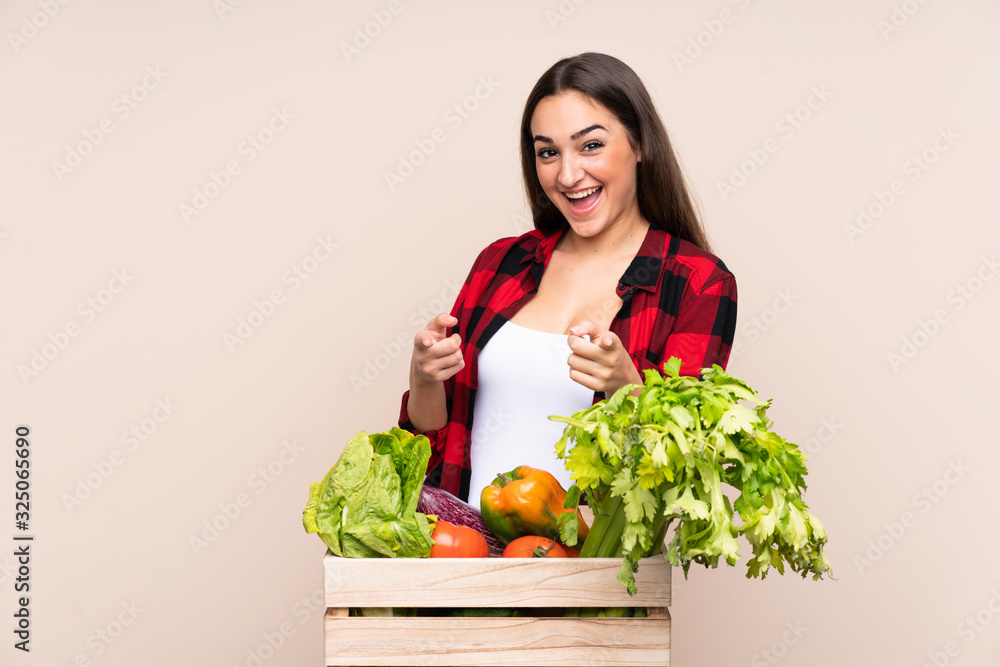 Farmer with freshly picked vegetables in a box isolated on beige background points finger at you Farmer with freshly picked vegetables in a box isolated on beige background points finger at you