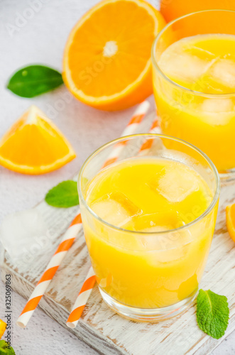 Freshly squeezed orange juice with ice in a glass with a straw on a wooden board on a light background with fresh oranges. Vertical orientation, close up.