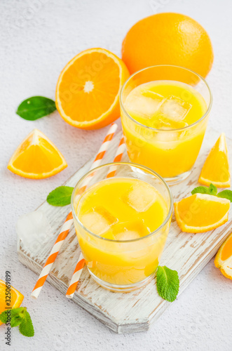 Freshly squeezed orange juice with ice in a glass with a straw on a wooden board on a light background with fresh oranges. Vertical orientation, close up.