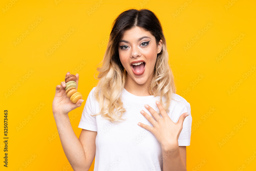 Teenager girl isolated on yellow background holding colorful French macarons with surprise expression Teenager girl isolated on yellow background holding colorful French macarons with surprise expression