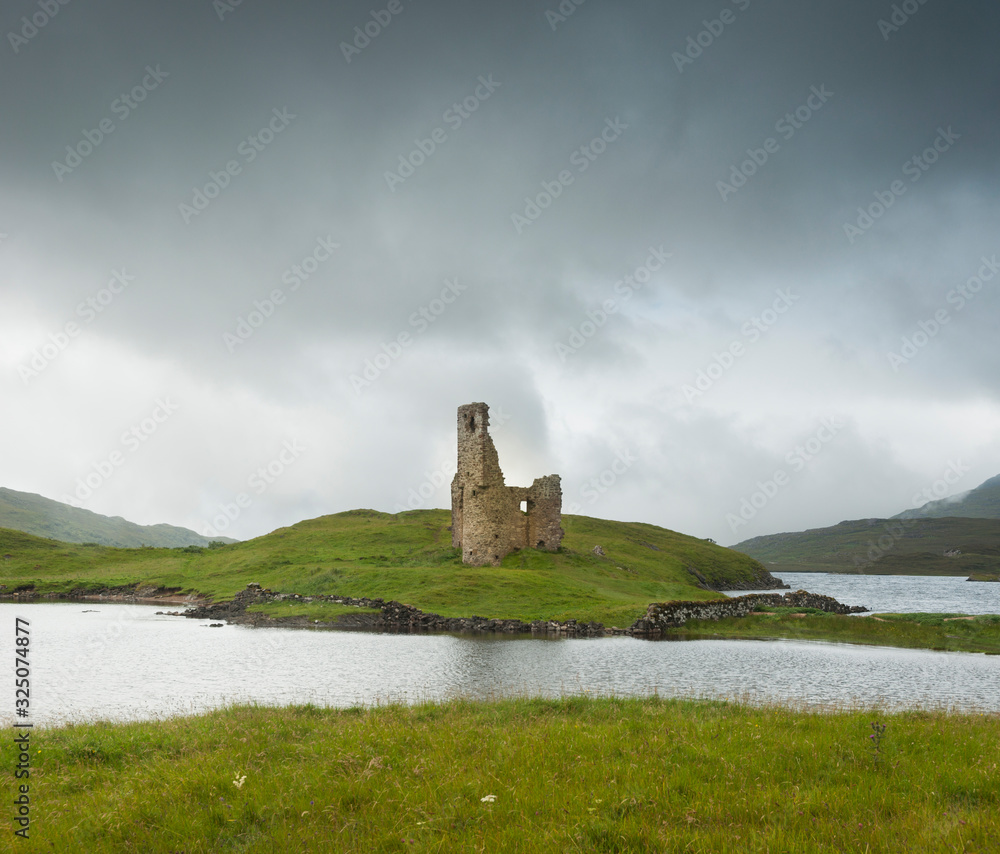 Landscape with ancient ruins of the small castle of Ardvreck in Scotland on a lake and with cloudy skies.