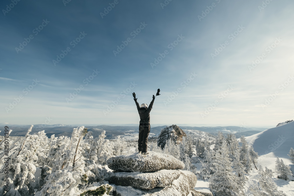 Hiker female standing with hands up achieving cliff top, admiring winter mountain landscape. Happy freerider snowboarder tourist in winter