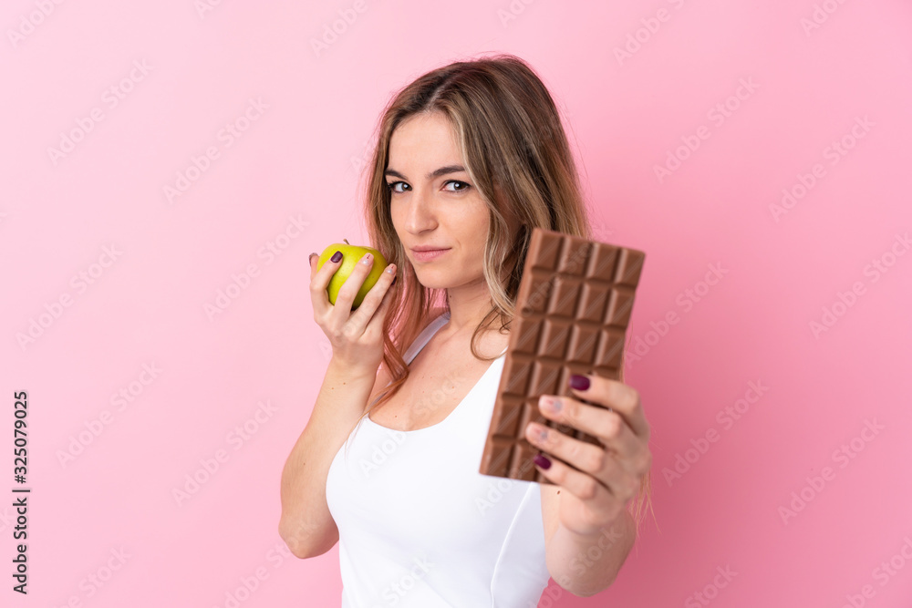 Young woman over isolated pink background taking a chocolate tablet in one hand and an apple in the other Young woman over isolated pink background taking a chocolate tablet in one hand and an apple in the other