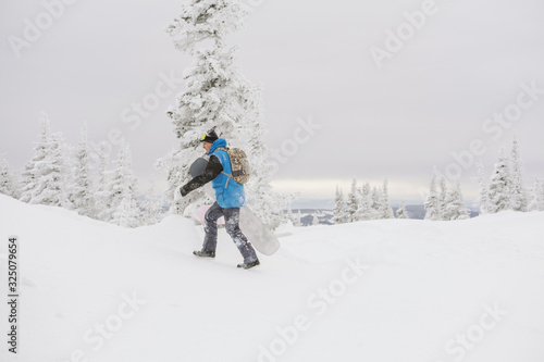 Snowboarder free rider man walking on snowy slop, snow covered trees on background, bad weather, siberian winter snow powder day in ski resort