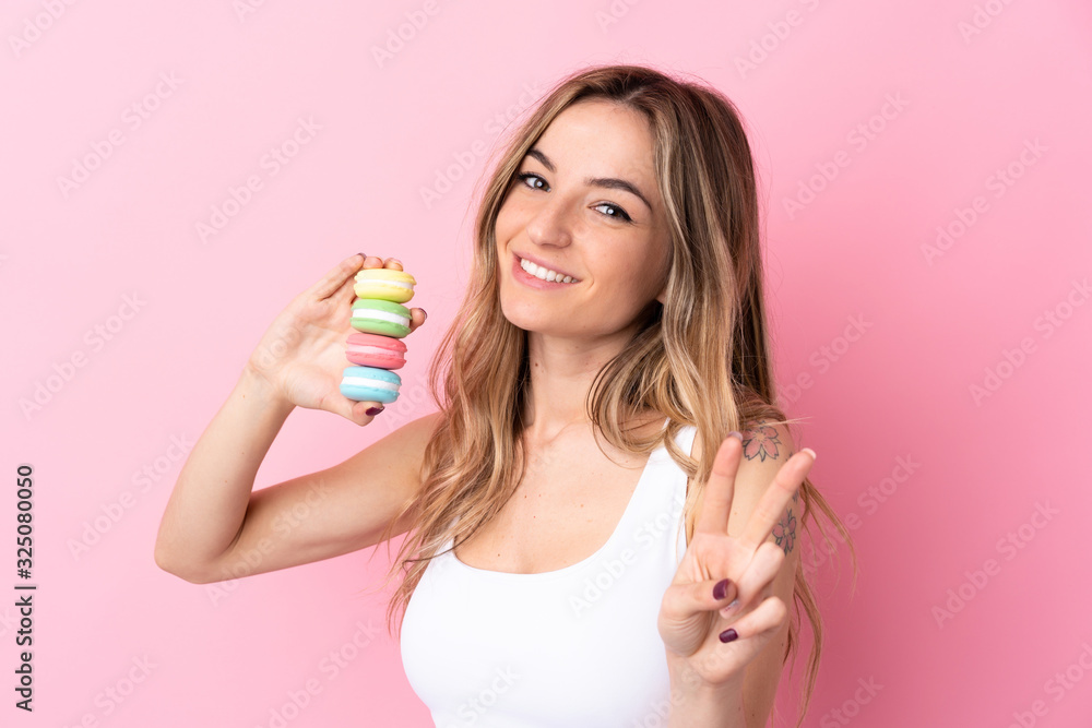 Young woman with macarons over isolated pink background smiling and showing victory sign Young woman with macarons over isolated pink background smiling and showing victory sign