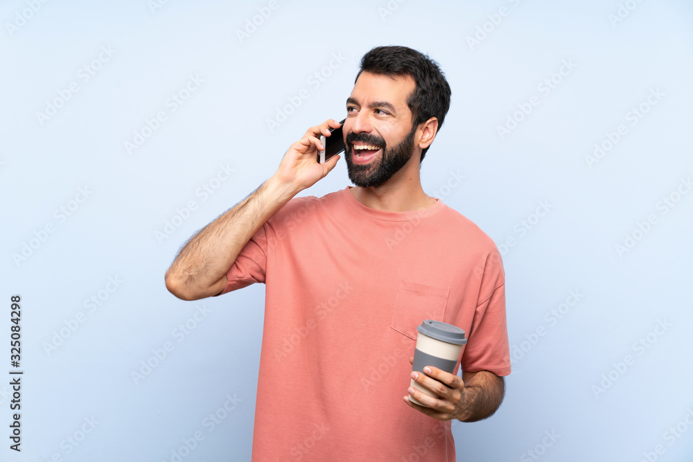 Young man with beard holding a take away coffee over isolated blue background keeping a conversation with the mobile phone Young man with beard holding a take away coffee over isolated blue background keeping a conversation with the mobile phone