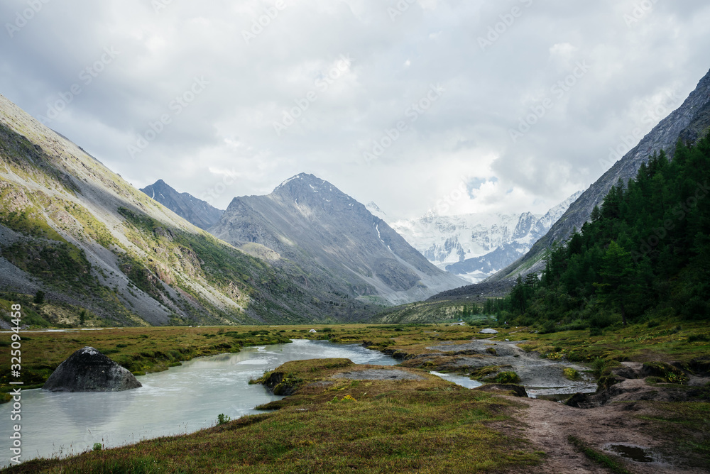 Awesome alpine view to mountain lake and great glaciers under gloomy sky. Dark atmospheric highland scenery with high snowy mountains and big rocks. Wonderful mountain landscape in cloudy weather.