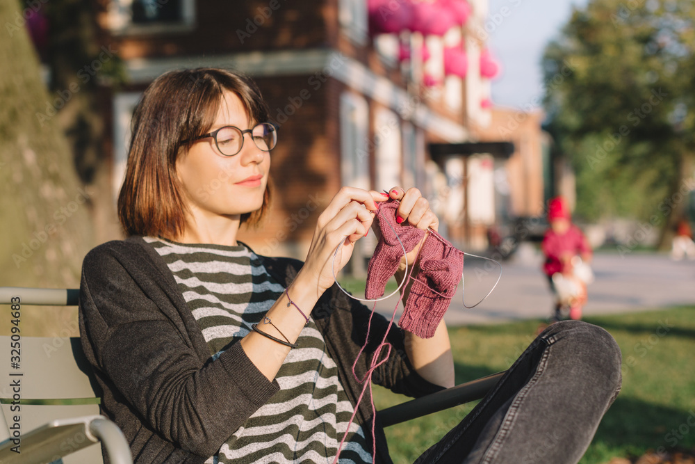 Obraz premium Close up photo of cute pretty girl in glasses and striped t-shirt. Attractive woman sitting in chair and knitting socks from purple yarn outdoor. Needleworks and hobby concept.