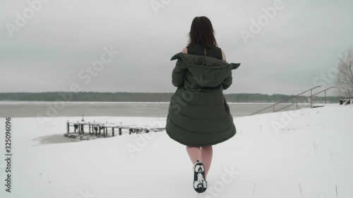 Back view of woman walking on snow takes her coat off and moves ahead to pond covered with ice and snow. Slow motion.