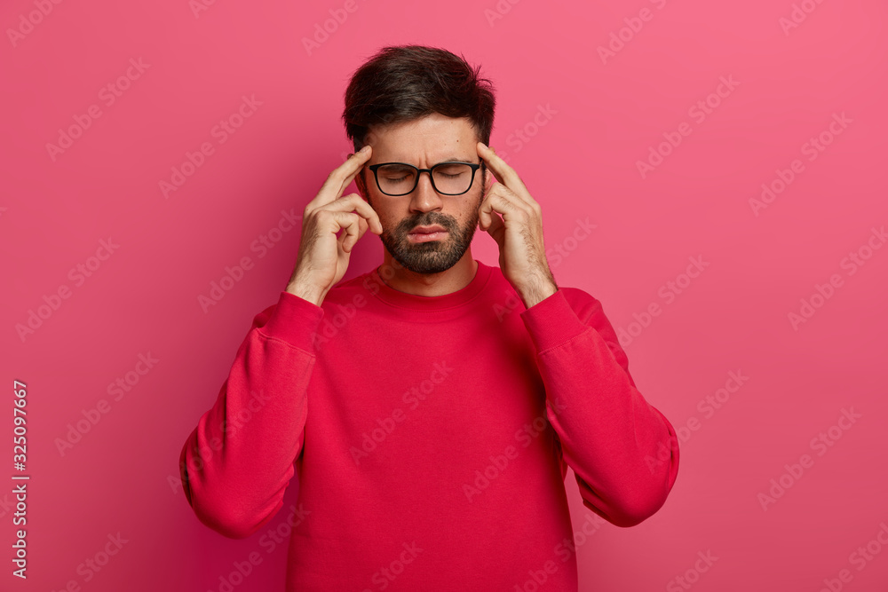 Stressed unshaven man closes eyes, keeps index fingers on temples, feels strong migraine, tries to concentrate on something, dressed in bright red sweater, isolated on rosy background. Health problems Stressed unshaven man closes eyes, keeps index fingers on temples, feels strong migraine, tries to concentrate on something, dressed in bright red sweater, isolated on rosy background. Health problems