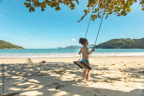 Beautiful girl swinging on wooden swing on the tree at the beach, happy smiling girl with red hair, playing on the beach on vacation trip, at Parnaioca beach, in Ilha Grande, in Rio de Janeiro, Brazil