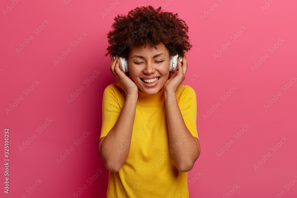 People and modern technologies concept. Smiling dark skinned woman enjoys melody, touches headphones, keeps eyes closed, smiles pleasantly, wears yellow t shirt, isolated on pink studio wall People and modern technologies concept. Smiling dark skinned woman enjoys melody, touches headphones, keeps eyes closed, smiles pleasantly, wears yellow t shirt, isolated on pink studio wall