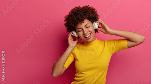 Overjoyed African American woman listens modern music via wireless headphones, enjoys good sound and chills with eyes closed, smiles broadly, wears casual yellow t shirt, isolated on pink wall