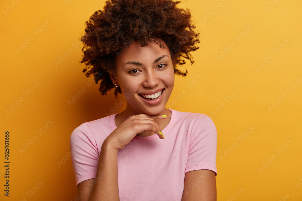 Portrait of optimistic friendly looking woman holds hand under chin, looks with charming exression at camera, has fun, expresses positive good emotions, isolated on yellow wall, dressed casually Portrait of optimistic friendly looking woman holds hand under chin, looks with charming exression at camera, has fun, expresses positive good emotions, isolated on yellow wall, dressed casually