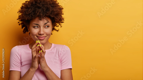 Horizontal shot of pretty woman with cunning expression steepls fingers and looks with intention aside, thinks about something mysterious, has evil plan, wears casual t shirt, isolated on yellow wall