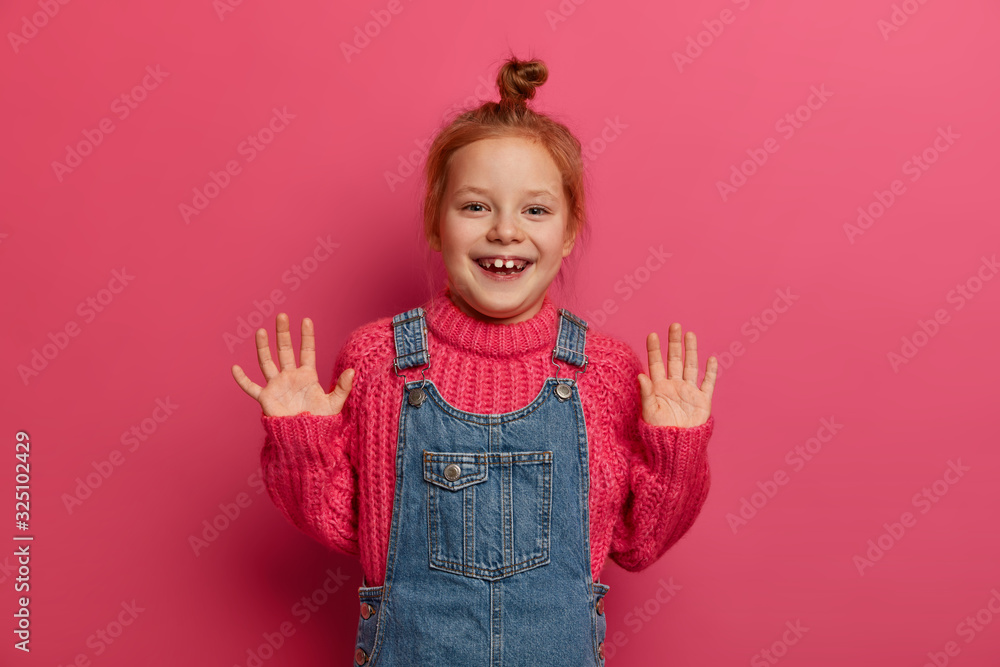 Playful positive girl with ginger hair combed in bun, raises palms and has good mood, poses for family photo in studio, wears knitted sweater and sarafan, has joyful expression isolated over rosy wall Playful positive girl with ginger hair combed in bun, raises palms and has good mood, poses for family photo in studio, wears knitted sweater and sarafan, has joyful expression isolated over rosy wall