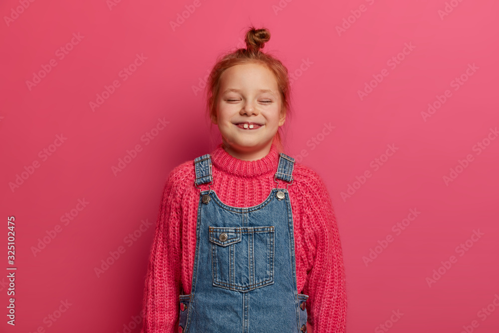 Small five years old girl with ginger hair bun and two stick out teeth, wears warm knitted sweater and denim sarafan, laughs from joy, keeps eyes closed, watches funny cartoon, isolated on pink wall Small five years old girl with ginger hair bun and two stick out teeth, wears warm knitted sweater and denim sarafan, laughs from joy, keeps eyes closed, watches funny cartoon, isolated on pink wall