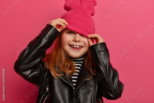 Cropped image of little girl looks from hat, hides face, wears stylish black leather jacket, dressed in fashionable clothes has positive ambitious look isolated on pink wall. Children, emotions, style