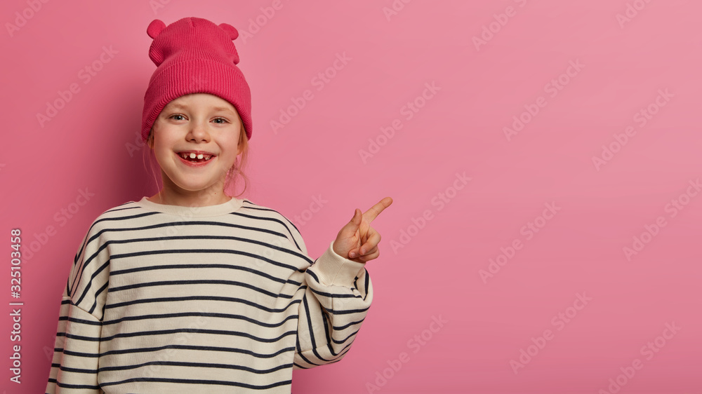 Adorable preschooler feels happy while points at upper right corner, demonstrates something interesting, attracts your attention to discounts in toy shop, wears rosy hat and striped sweater. Adorable preschooler feels happy while points at upper right corner, demonstrates something interesting, attracts your attention to discounts in toy shop, wears rosy hat and striped sweater.