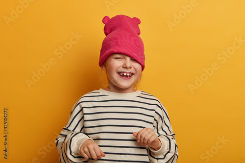 Sincere emotional child plays with new hat, dressed in striped jumper, laughs and cheers something, has funny joyful expression, playful mood, going crazy, isolated on vibrant yellow background