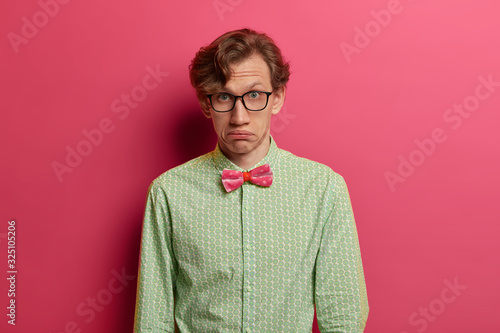 Fotografie, Obraz Indoor shot of embarrassed surprised man looks doubtfully, purses lips, wears spectacles, formal shirt and pink bowtie, looks direcly at camera, isolated over rosy background