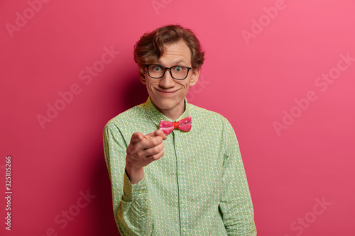 Pleasant looking cheerful European male nerd indicates at camera, has funny look, smiles broadly, wears shirt and spectacles, chooses you, isolated on pink studio wall. People, choice concept