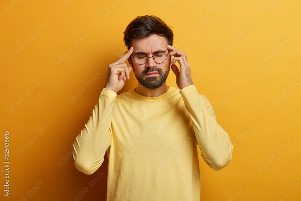 Overworked frustrated bearded man massages temples, suffers from severe migraine, closes eyes to relieve pain, wears optical glasses and casual yellow sweater, stands in studio, tries to calm down Overworked frustrated bearded man massages temples, suffers from severe migraine, closes eyes to relieve pain, wears optical glasses and casual yellow sweater, stands in studio, tries to calm down