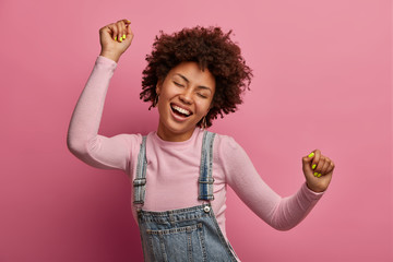 Happy dark skinned girl enjoys every moment of life, dances and moves, raises arms and clenches fists, closes eyes, has good mood, wears denim sarafan and turtleneck, isolated on pink background