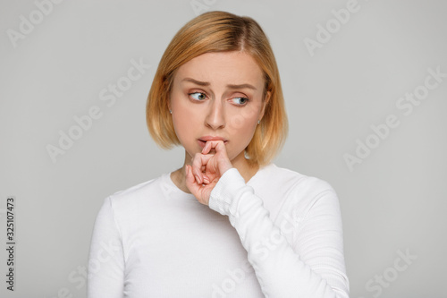 Oops, how awkward! Close up portrait of shy awkward young woman biting lips feeling embarrassed, confused and nervous, looking aside, covers mouth with her finger. Isolated on grey background. 
