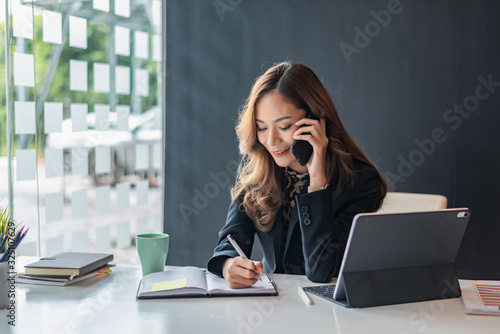 Happy young asian woman talking on the mobile phone and smiling while sitting at her working place in office. photo