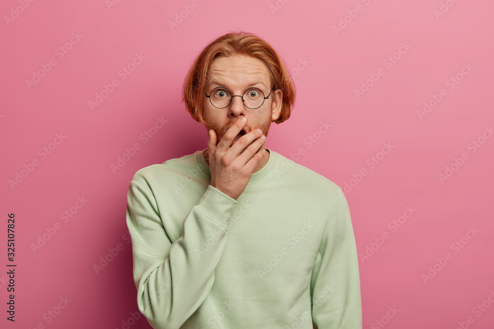 Isolated shot of stunned bearded red haired hipster man covers opened mouth, cannot believe in shocking news, sees something unbelievable, dressed in casual jumper, poses against pink background Isolated shot of stunned bearded red haired hipster man covers opened mouth, cannot believe in shocking news, sees something unbelievable, dressed in casual jumper, poses against pink background