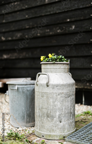 A traditional milkcan with yellow violet and an old trashcan behind it against a black wooden barn wall