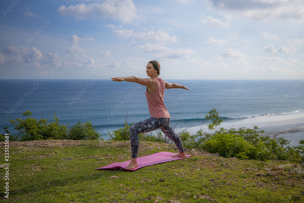 Young woman practicing yoga, standing in Virabhadrasana pose. Outdoor yoga on the cliff. Warrior pose. Bali, Indonesia Young woman practicing yoga, standing in Virabhadrasana pose. Outdoor yoga on the cliff. Warrior pose. Bali, Indonesia