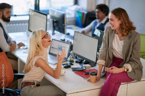  female colleagues talking and smiling at work in modern open space office, with male colleagues in the background. casual, open space, modern, office, business concept
