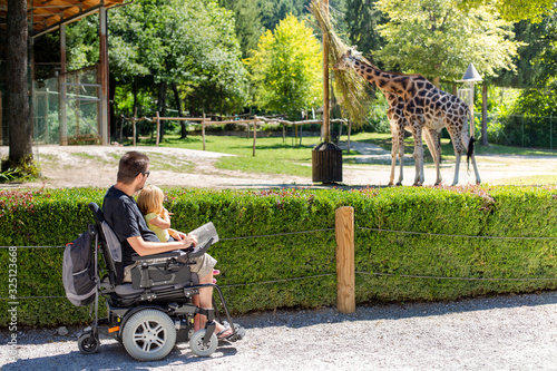 Disabled Father in Wheelchair enjoying Nature with Daughter Girl in Outside zoo park on a sunny day.