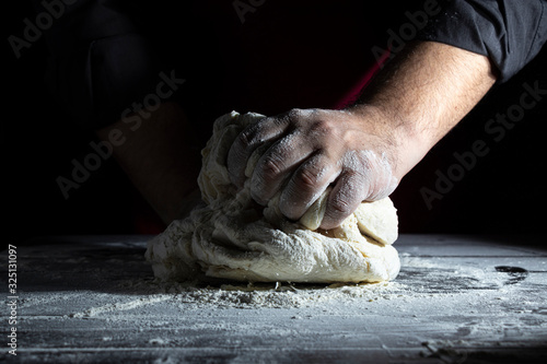 Male chef kneads dough on a wooden table. Dark background. Close-up.