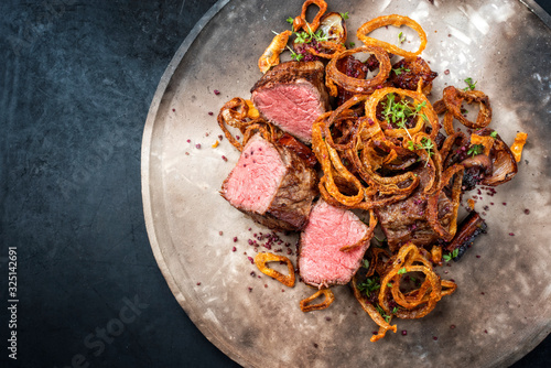 Traditional dry aged sliced beef tenderloin with fried onion rings and potatoes served as top view on a modern design plate with copy space left