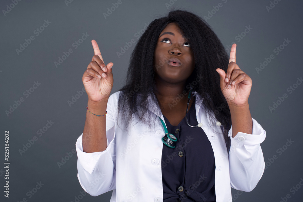 Portrait of beautiful elegant African American doctor woman over isolated background being amazed and surprised looking and pointing up with fingers showing something strange. Portrait of beautiful elegant African American doctor woman over isolated background being amazed and surprised looking and pointing up with fingers showing something strange.