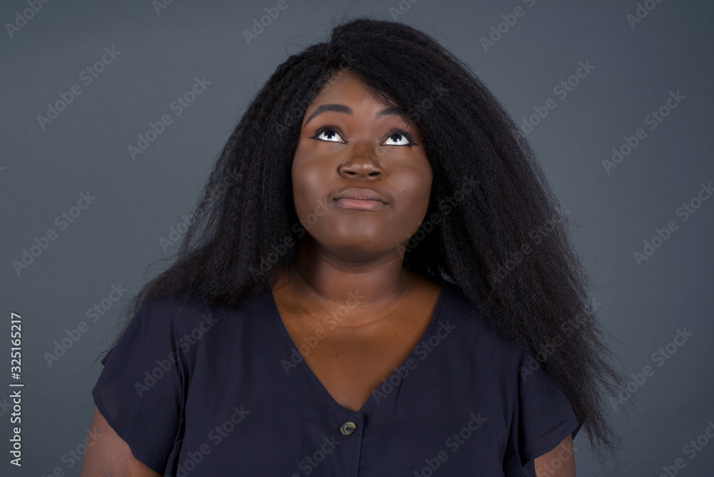 portrait of mysterious charming African American female with straight hair looking up with enigmatic smile. Beautiful smiling girl looking up standing against gray wall. portrait of mysterious charming African American female with straight hair looking up with enigmatic smile. Beautiful smiling girl looking up standing against gray wall.