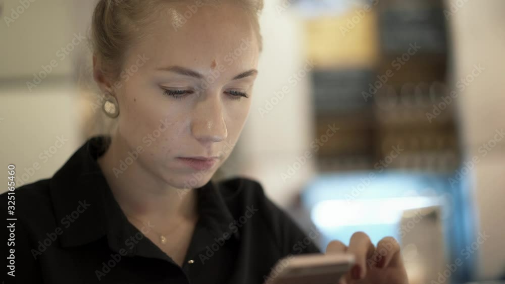 Handheld close up of serious female blonde hair holding a phone on blurred yellow background indoors. Close up face of blonde woman in black clothes typing in phone Handheld close up of serious female blonde hair holding a phone on blurred yellow background indoors. Close up face of blonde woman in black clothes typing in phone
