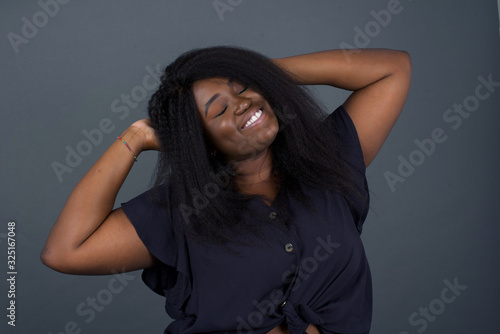 Confidence and coquettish concept. Portrait of charming young African American girl, smiling broadly with self-assured expression while holding hands over her head. Standing against gray background.