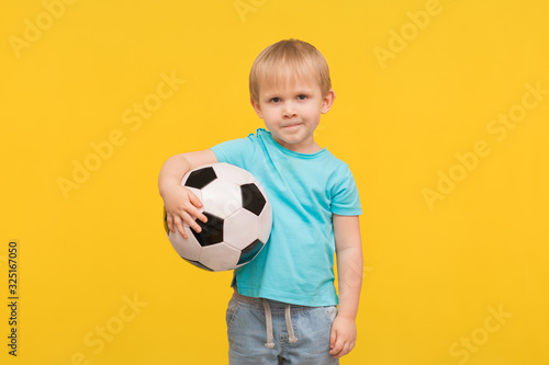 A charismatic handsome blond boy stands with a soccer ball in his hands and looks at the camera with a challenge and expectation on a yellow background.