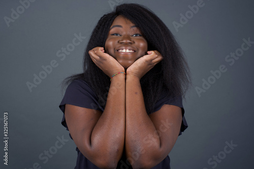 Portrait of young cute African American student being overwhelmed with emotions, expressing excitement and happiness with closed eyes and hands near face while smiling broadly over gray background.